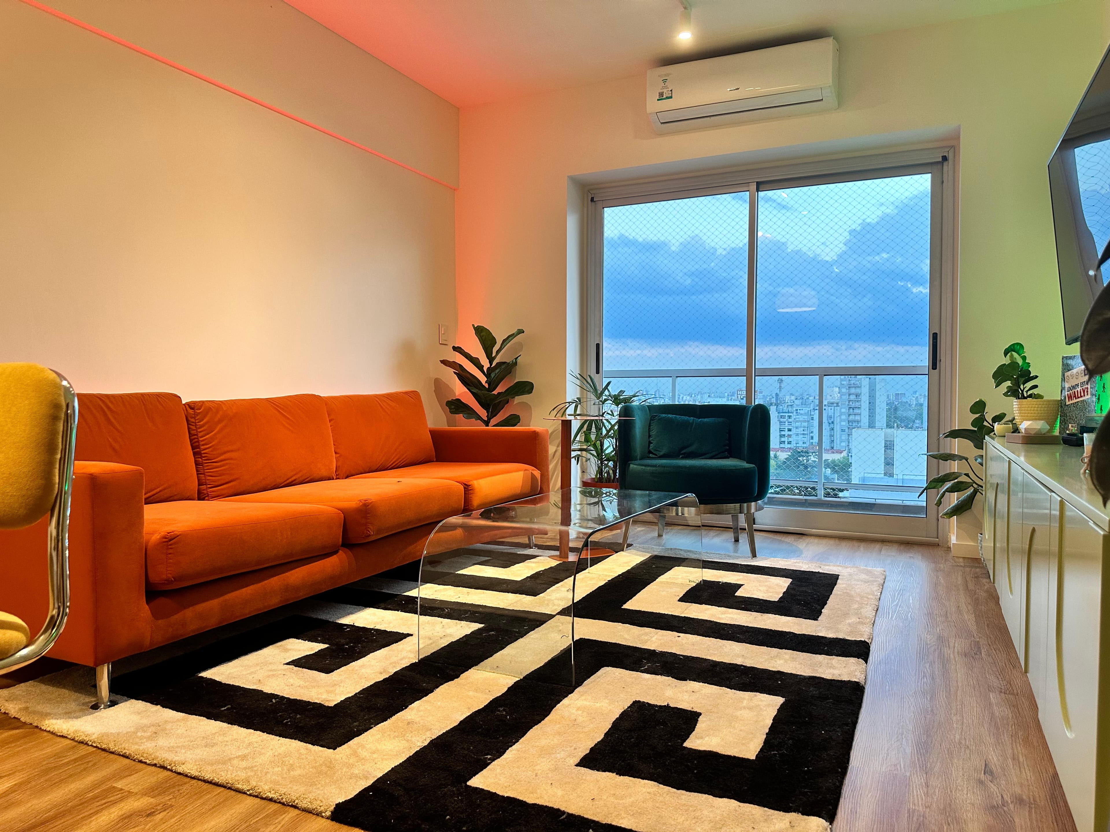 Living con sillón de terciopelo naranja, alfombra con greca blanca y negra, y ventanal con vista a la ciudad